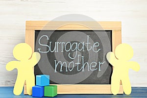 Small chalkboard with phrase Surrogate Mother, paper people cutouts and cubes on blue wooden table