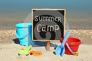 Small chalkboard with text SUMMER CAMP and beach toys on sand