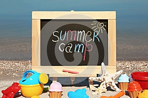 Small chalkboard with text SUMMER CAMP and beach toys on sand