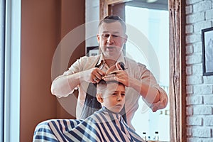 Small charming boy is getting trendy haircut from expirienced barber at fashionable hairdressing salon