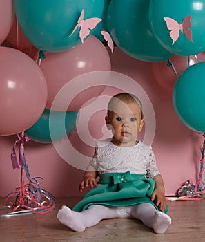 A small, charming child, a girl, celebrates her first birthday, sitting next to her with balloons, on a pink background. Children