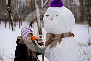 A small cheerful girl holds a big carrot, the nose of a big snowman