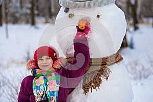 A small cheerful girl holds a big carrot, the nose of a big snowman