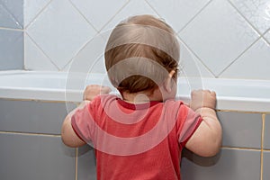 A small child in the bathroom looking into the bath, holding the edge