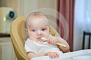 a small child with blue eyes is holding a spoon sitting in a high chair.