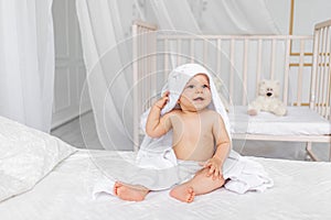 A small child a boy 8 months old is sitting in a towel on a white bed in a light nursery in diapers after bathing