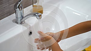 A small child boy opens the tap and washes his hands with soap under running water in sink. Clean your hands to prevent