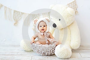 A small child a boy is sitting in a basket in a bear hat with a large Teddy bear