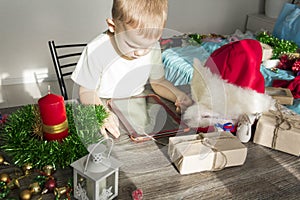 A small child with Christmas gifts is sitting at a New Year's wooden table with a tablet in his hands