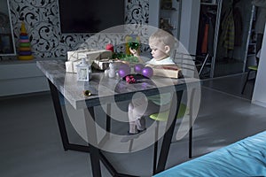A small child with Christmas gifts is sitting at a New Year\'s wooden table with a tablet in his hands
