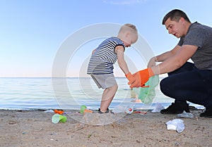 A small child collects trash on the beach. His dad points his finger where to throw garbage. Parents teach children cleanliness.