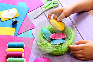 Small child does Easter decor. Child holds a felt egg in hand. Colorful felt Easter eggs and sisal nest, sewing tools and material