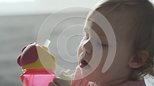 small child eat banana and drink water from yellow baby drinker. A funny baby in pink T-shirt with blond curly hair sits