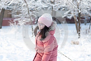 A small child in glasses, warm clothes, a knitted hat and mittens pulls a sled by the rope on the rolled snow. Photo shoot on a