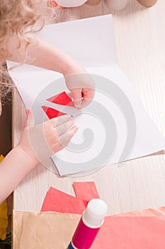 a small child glues paper, children`s safe scissors and colored paper on the table