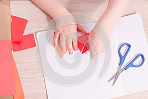 A small child glues paper, children`s safe scissors and colored paper on the table