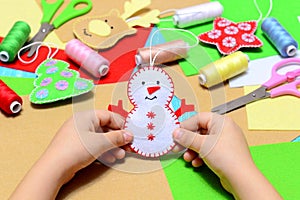 Small child holding a felt Christmas snowman in hands. Little kid shows Christmas ornament crafts. Workplace in kindergarten