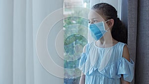 A small child or little girl in a protective medical mask looks out the window from behind a curtain, with a sad