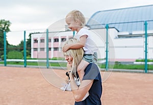 Small child, a little girl sits on the shoulders of mother smiling, laughing, playing, having fun at tennis court.