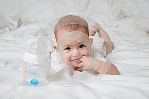 A small child is lying on a white bed on his stomach. The girl put two fingers into her mouth. Nearby there is a plastic bottle