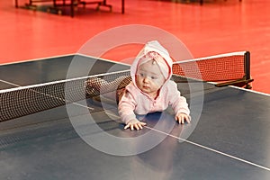 A small child in a pink suit is lying on his stomach on a table tennis table