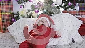 A small child in a red santa suit lying under the Christmas tree and smiles.