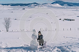 Small child sits in the snow and looks at his mother making a snowman. Back view