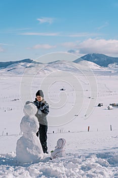 Small child sits in the snow and watches his mother build a snowman in a mountain valley. Back view