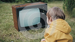 Small child is sitting on grass and watching an old retro TV. TV is strobing