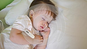 Small child sleeps in hospital ward on white bedding and measures temperature with thermometer. Treatment of children in