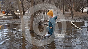 A small child walks in a large puddle with a stick in his hand in the spring
