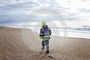 A small child in warm clothes walks along the seashore during the cold season. Activity and walks. Back view