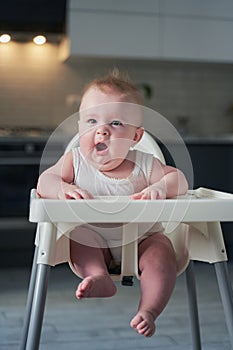 A small child in a white bodysuit sits in a high chair and yawns