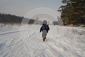 Small child in a winter suit and scarf runs into the distance along a snow-covered road next to the forest, view from