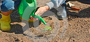 A small child in yellow boots helps his mother water the plants in the garden