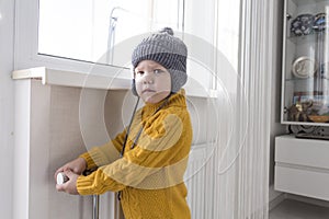 A small child in a yellow sweater and a gray hat is standing near a heater with a thermostat.