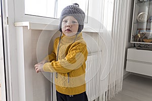 A small child in a yellow sweater and a gray hat is standing near a heater with a thermostat.