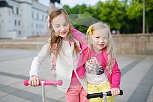 Small children learning to ride scooters in a city park on sunny summer evening. Cute little girls riding rollers.