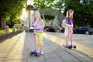 Small children learning to ride scooters in a city park on sunny summer evening. Cute little girls riding rollers.