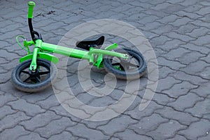 A small childrenâ€™s two-wheeled bicycle of green color lies on its side on a street tile of gray color without people and nobody