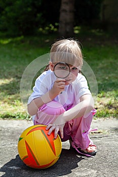 Small clever happy girl, child looking through a magnifying glass, kid with a loupe and a basketball playing in the garden outside