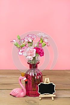 Small colored pink carnations in vase, frame, figure of flamingo on wooden table and pink wall Hello spring, seasonal concept Mock
