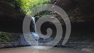 Small creek waterfall with water falling through the stone cave. Fast stream flowing down from the forest spring, through the hill