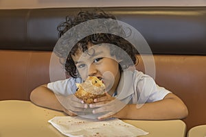 Small curly hair multiracial boy sitting in a coffee shop booth eating a muffin