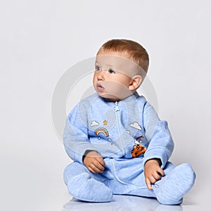 Small cute baby boy in blue warm cotton comfortable jumpsuit sitting on floor and looking aside