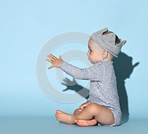 Small cute baby boy in grey comfortable jumpsuit, decorative crown on head and barefoot sitting and reaching for something