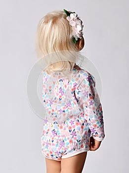 Small cute blonde baby girl in floral jumpsuit with flower decoration in hair standing backwards