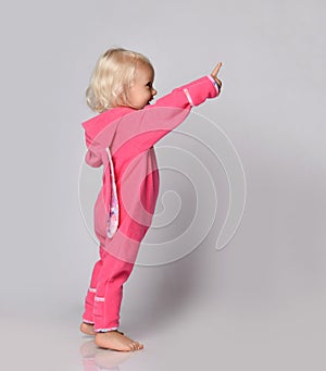 Small cute blonde positive baby girl in pink warm comfortable jumpsuit standing and pointing up with finger