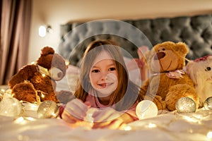 A small cute girl is lying alone on her stomack on a big bed barefoot having a good time looking into a camera, smiling