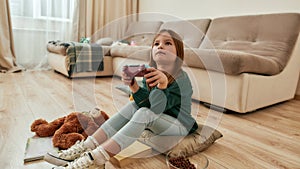 A small cute girl watches attentively at a screen of a TV playing videogame holding a gamepad while sitting on a floor